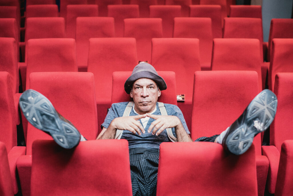 Peter Shub con sombrero, sentado entre las butacas rojas de un teatro.