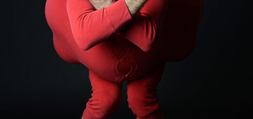 Eric Davis como Red Bastard, vestido de rojo, con rostro maquillado de blanco y brazos cruzados frente a un fondo negro.
