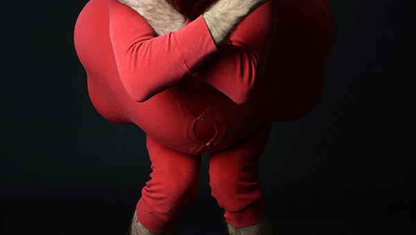 Eric Davis como Red Bastard, vestido de rojo, con rostro maquillado de blanco y brazos cruzados frente a un fondo negro.