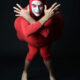 Eric Davis como Red Bastard, vestido de rojo, con rostro maquillado de blanco y brazos cruzados frente a un fondo negro.